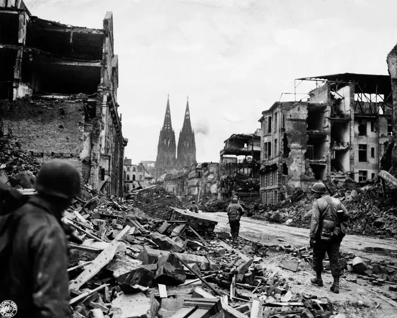 مدينة كولونيا بعد الحرب العالمية الثانية، تظهر جنودًا يسيرون بين الأنقاض مع كاتدرائية كولونيا في الخلفية، تعكس آثار القصف المدمر.