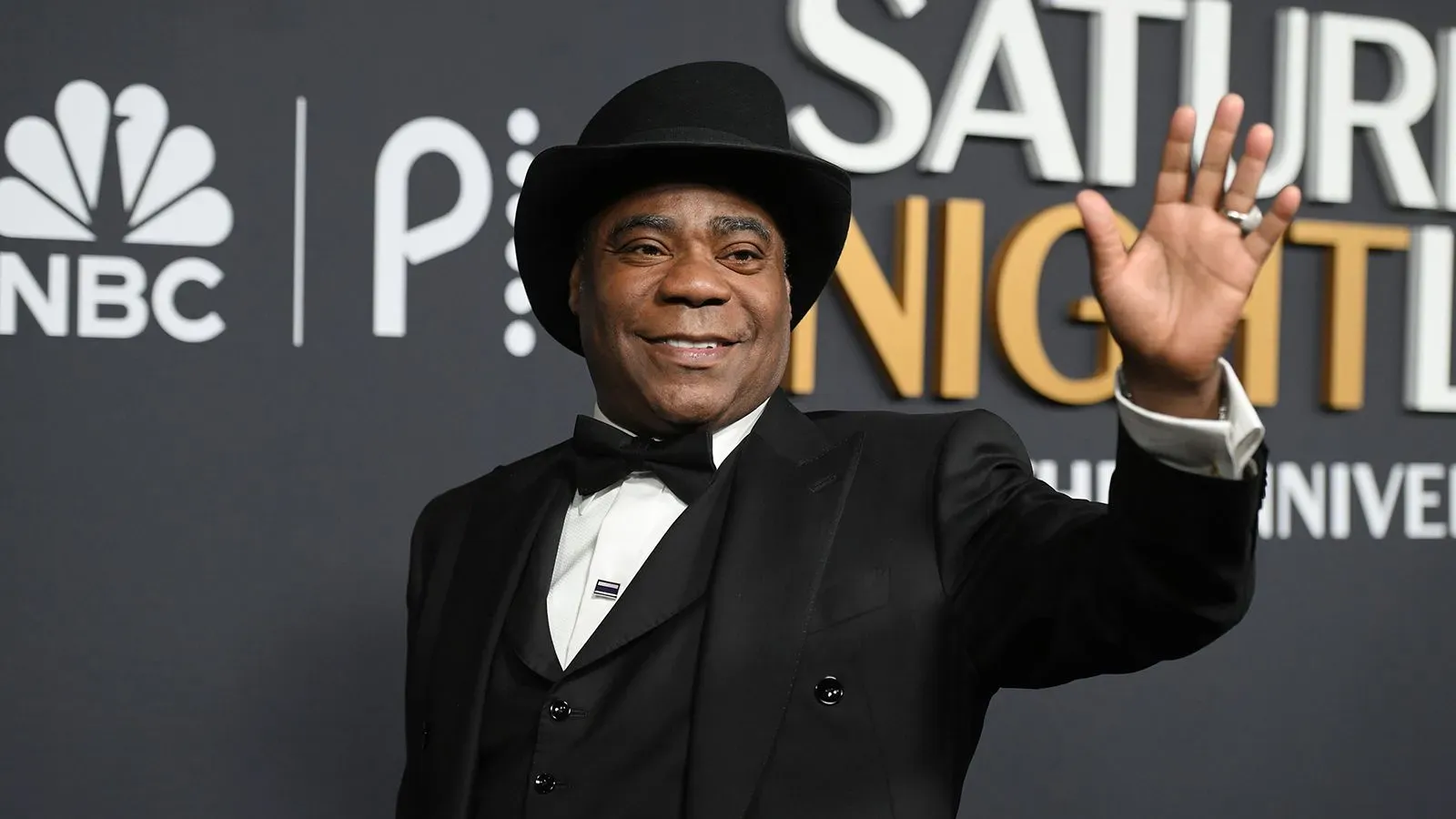 تريسي مورغان، الممثل الكوميدي المعروف، يرتدي بدلة أنيقة وقبعة، ويبتسم بينما يلوح بيده في حدث رسمي.