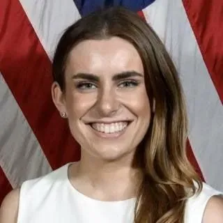 صورة تظهر تشامبرلين هاريس، العضو الجديد في لجنة الفنون الجميلة، مبتسمة أمام علم أمريكي، مما يعكس التوجهات السياسية في تجديد الجناح الشرقي.