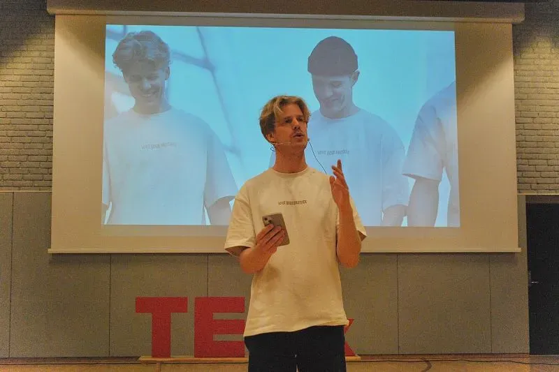 رجل يتحدث في حدث TED، مع عرض خلفي لصور لرجال آخرين. يركز على أهمية صداقات الرجال وتعزيز الروابط العميقة.