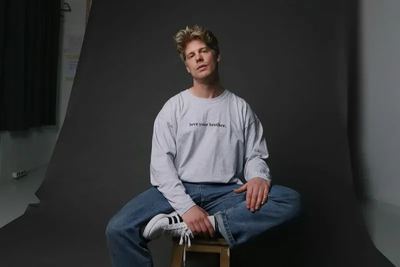 شاب يجلس على كرسي، يرتدي قميصًا مكتوبًا عليه "أحب أخاك"، مع خلفية داكنة، يعكس أهمية الصداقات بين الرجال.