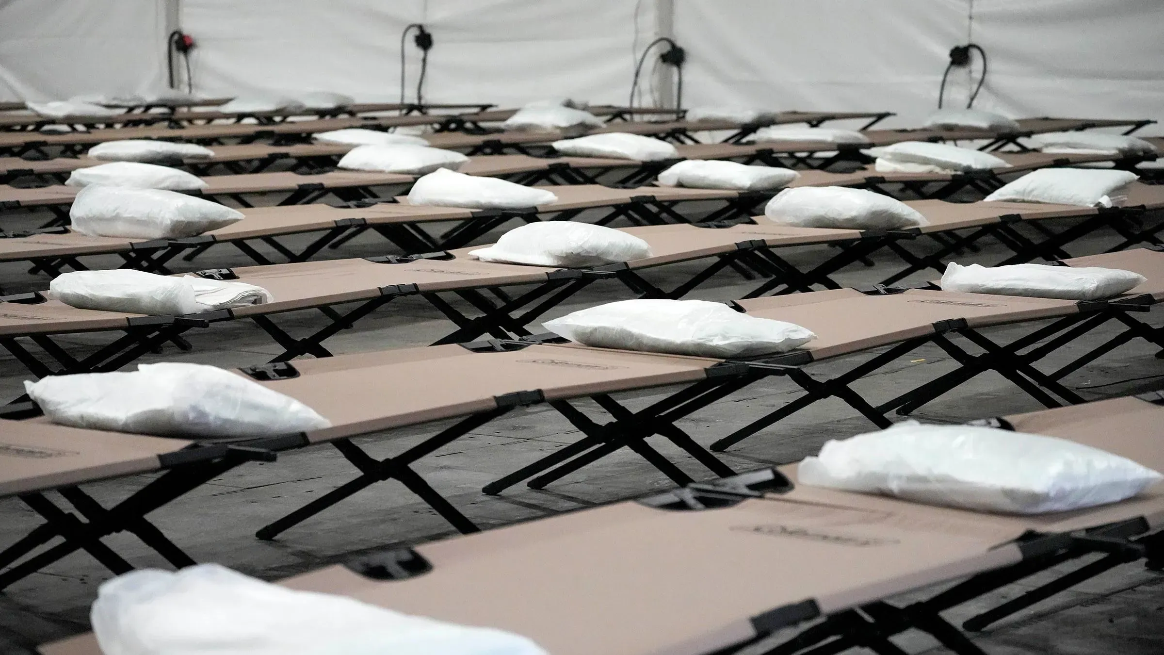 صفوف من أسرة مؤقتة مزودة بوسائد داخل خيمة، تعكس جهود توفير المأوى للمهاجرين في ظل مناقشات حول إعادة تخصيص التمويل.