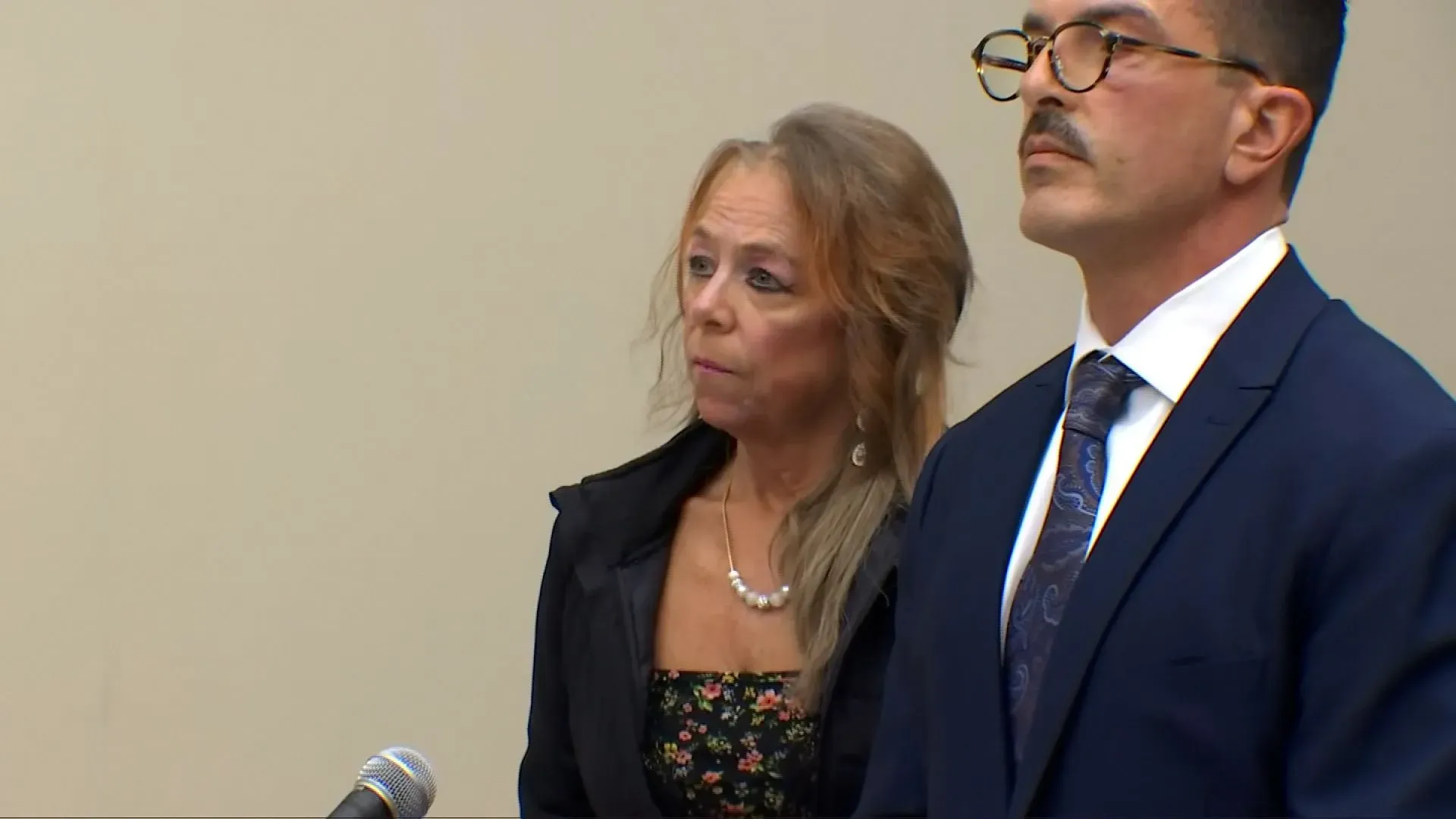 امرأة في قاعة المحكمة، ترتدي زيًا أسود مزينًا بالزهور، تقف بجانب محاميها خلال جلسة استماع بشأن اتهامات خطف وإساءة معاملة.