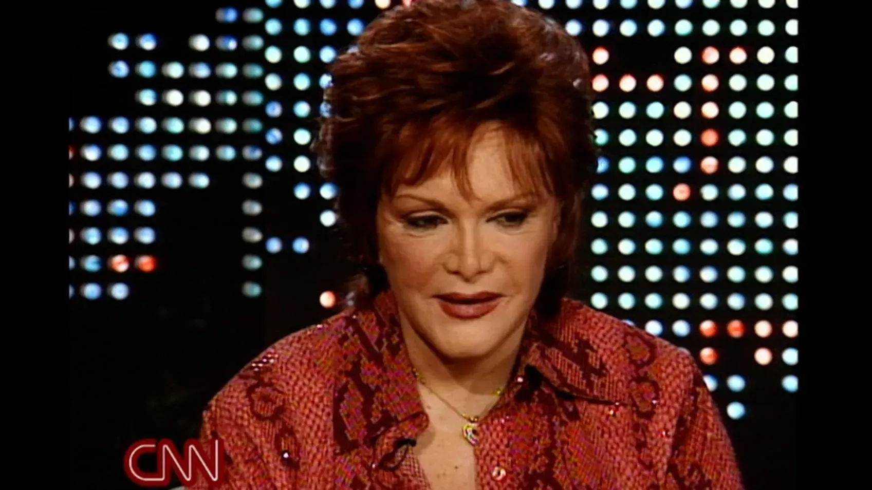 كوني فرانسيس، مغنية بوب شهيرة، تتحدث في مقابلة، مرتدية قميصاً مزخرفاً، مع خلفية مضيئة، تعكس مشوارها الفني وتأثيرها على الأجيال.