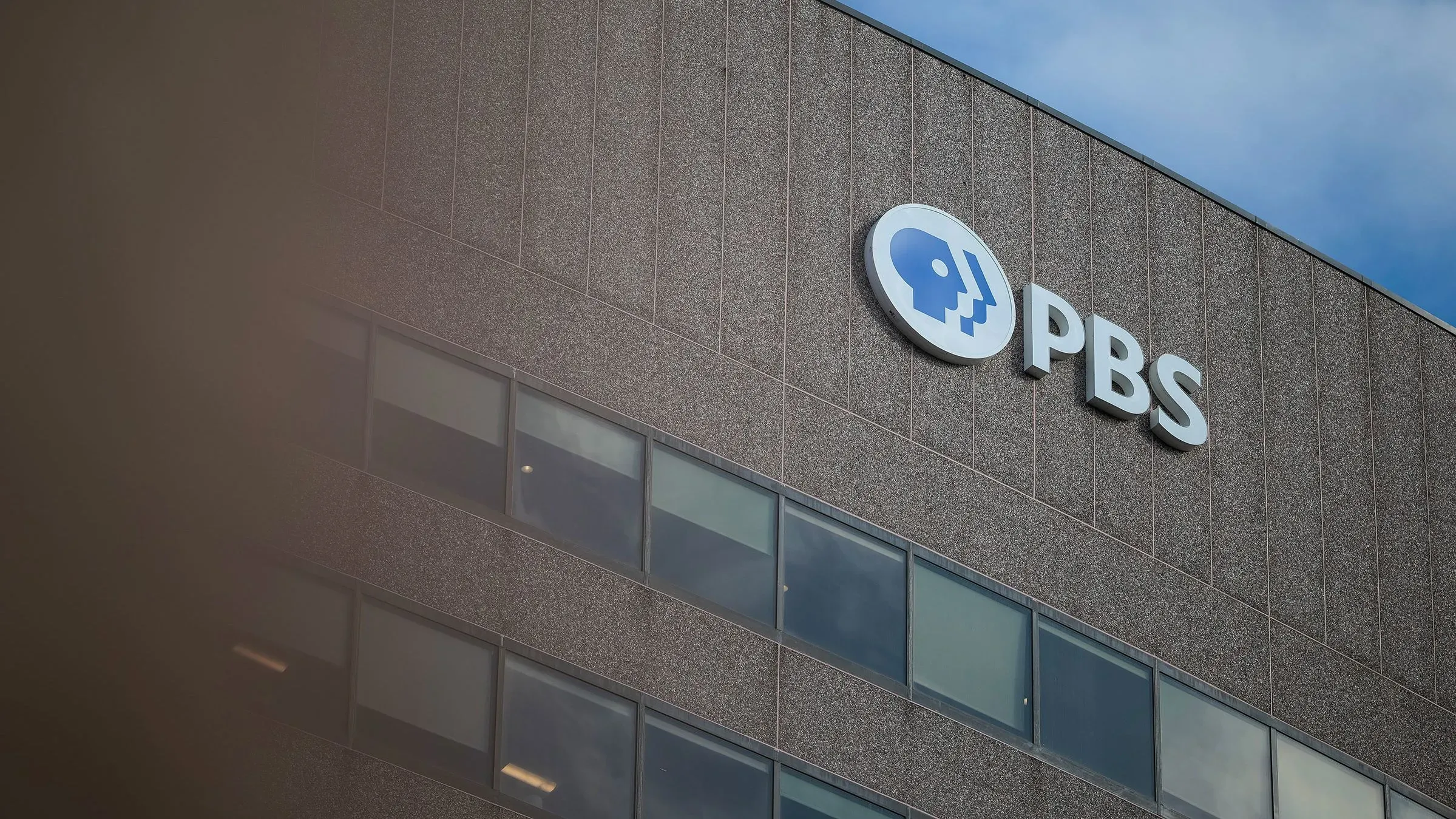شعار شبكة PBS على واجهة مبنى، يُظهر التوتر بين دعم البث العام والجهود السياسية لتخفيض التمويل الفيدرالي له.