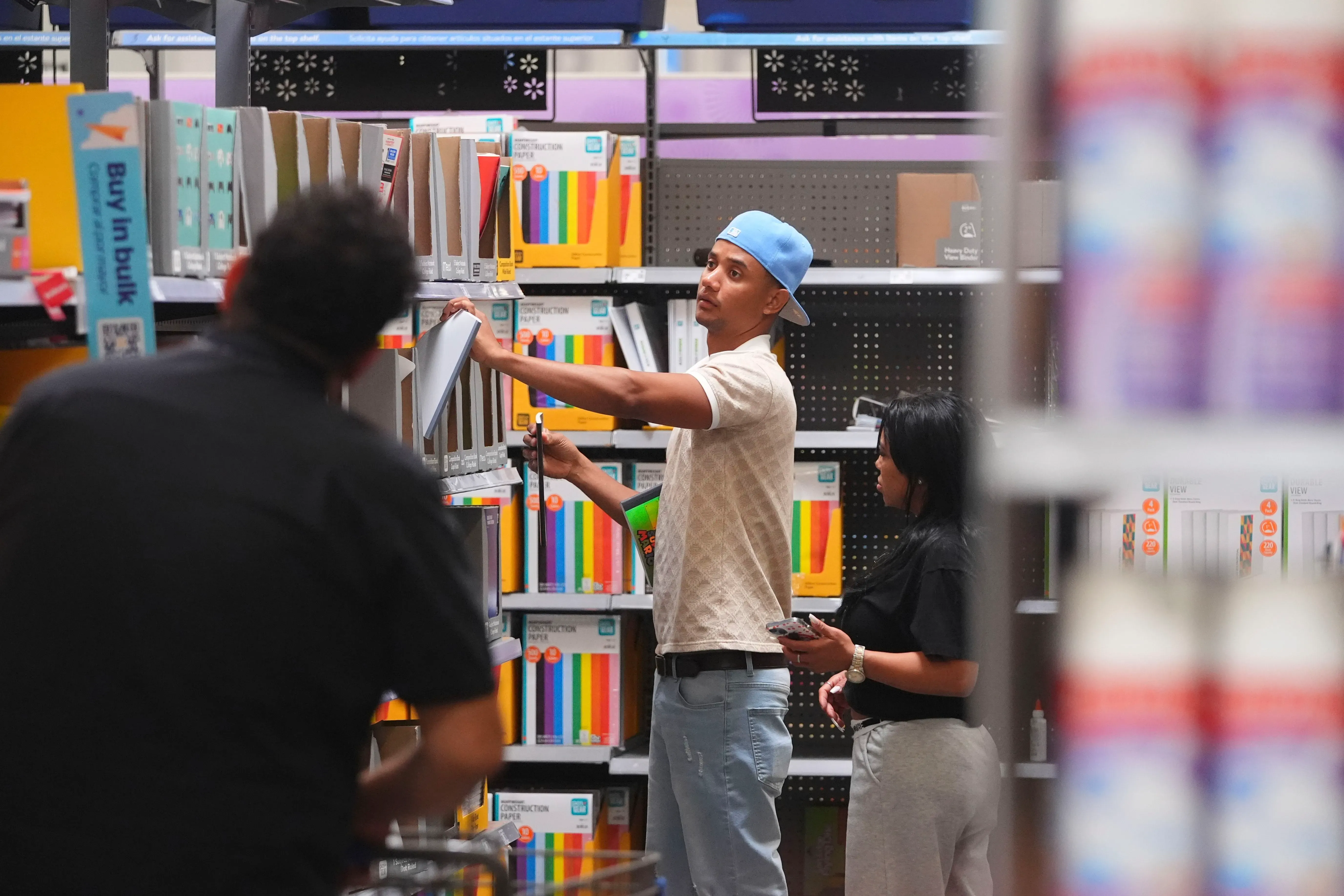 شخصان يتسوقان في متجر لبيع اللوازم المدرسية، حيث يقوم أحدهما بترتيب الأرفف بينما تحمل الأخرى هاتفها.