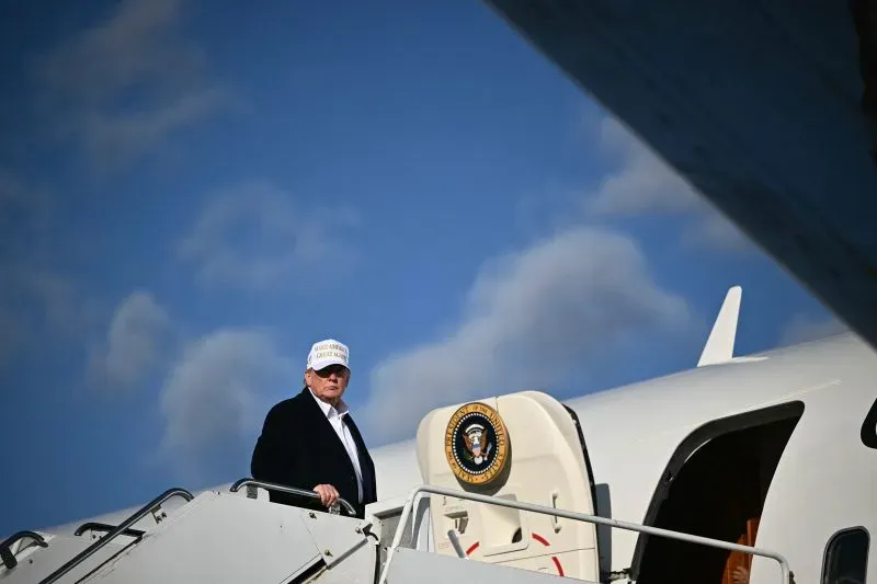 دونالد ترامب يصعد إلى طائرة رئاسية، مع شعار "اجعل أمريكا عظيمة مرة أخرى" على قبعة. الصورة تعكس التحديات الأمنية المتعلقة بالطائرة القطرية المقترحة.