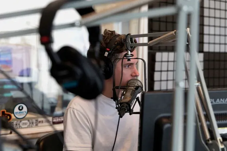 شخص يجلس أمام ميكروفون في استوديو إذاعي، مع سماعات على أذنه، يعكس تأثير خفض التمويل على الإعلام العام.