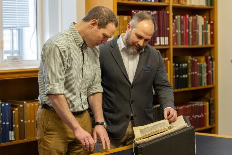 باحثان يتفحصان كتابًا قديمًا في مكتبة، محاطان برفوف تحتوي على كتب ومخطوطات تاريخية، في سياق دراسة أغنية وايد.