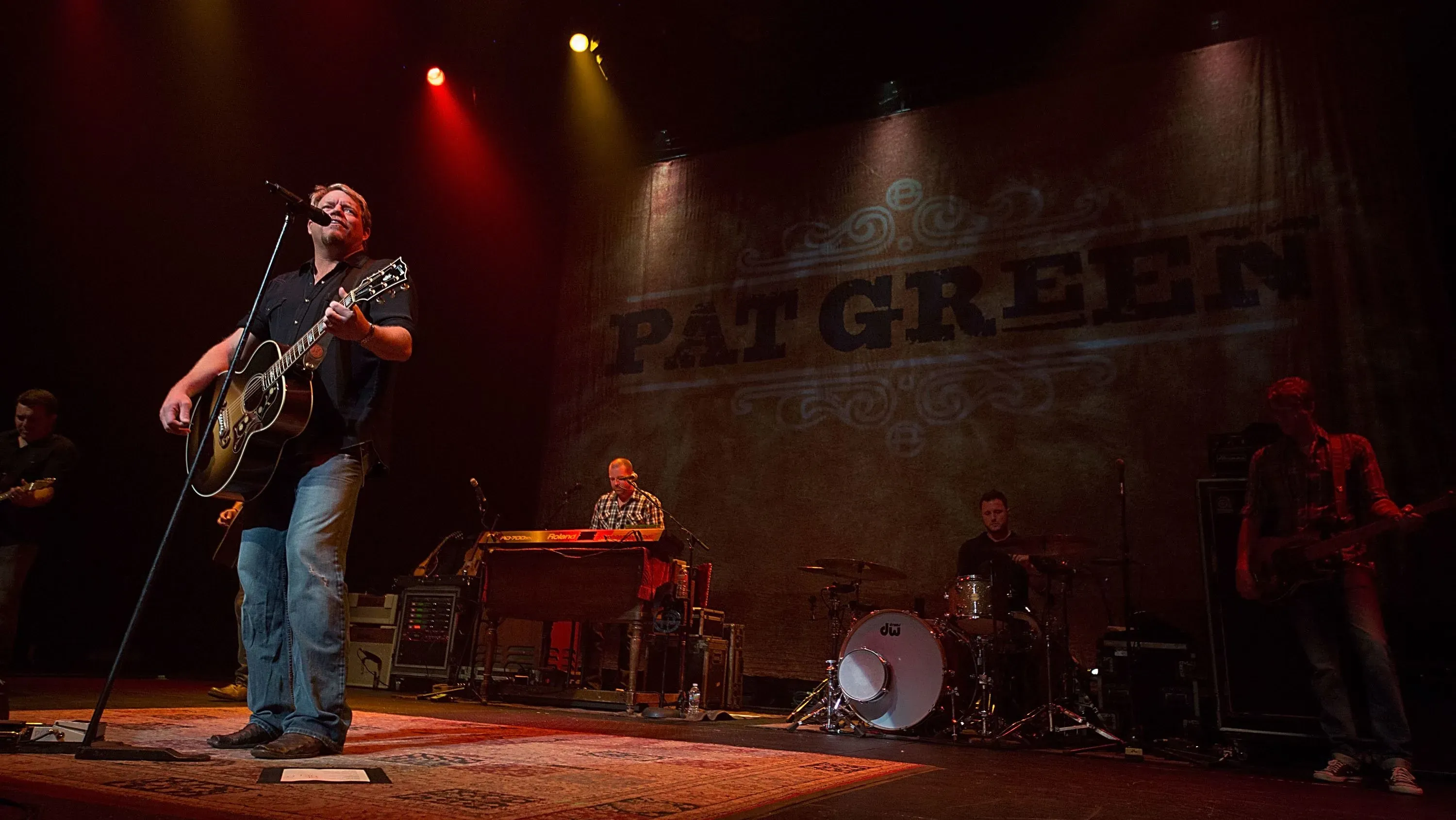 بات غرين يؤدي على المسرح خلال حفلة موسيقية، مع وجود فرقة موسيقية خلفه، في ظل أضواء ملونة، وسط أجواء حزينة بعد الفيضانات في تكساس.