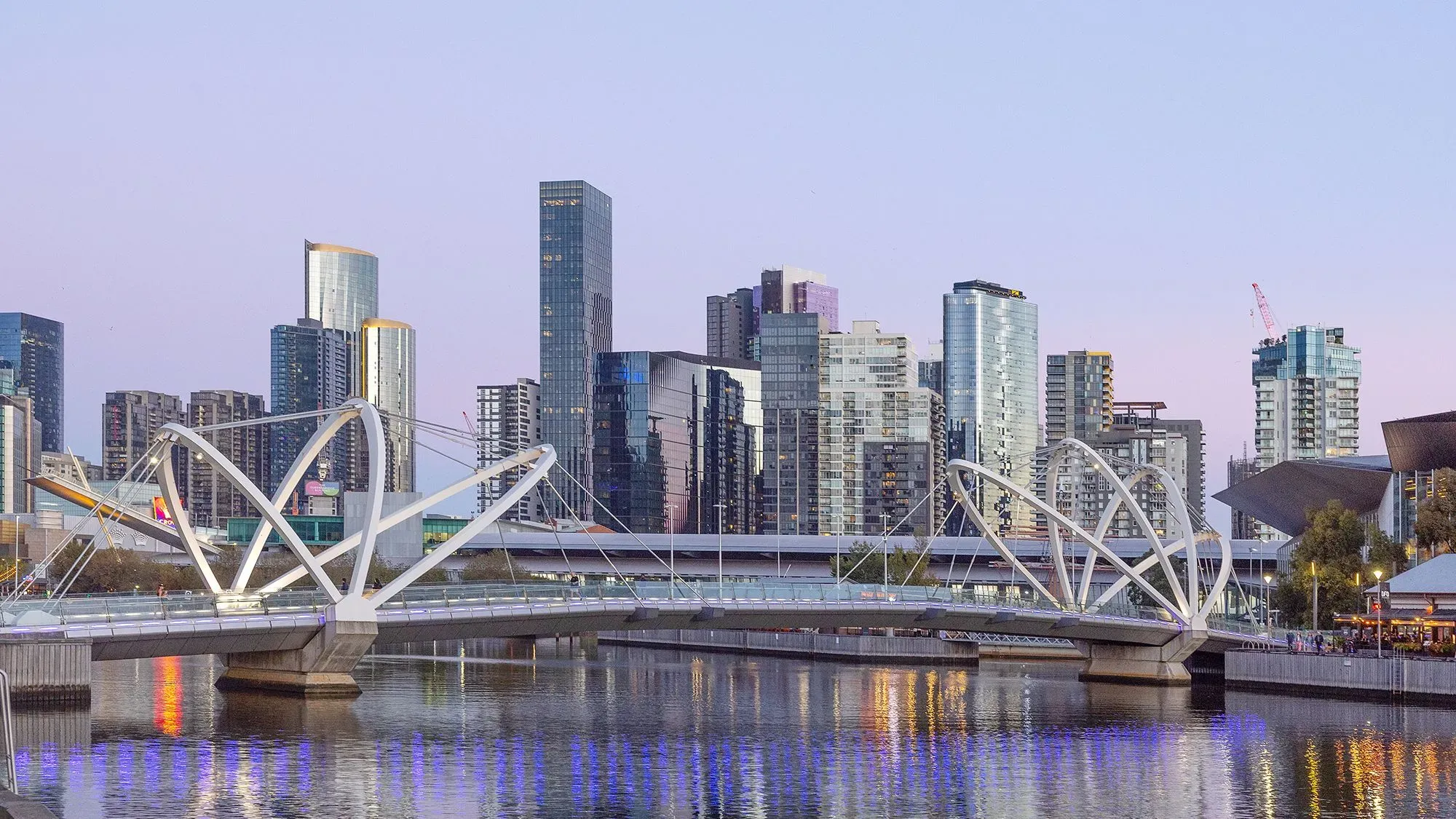 منظر لمدينة ملبورن مع نهر وجسر حديث، يظهر العمارة العصرية والمباني الشاهقة في خلفية الغروب.