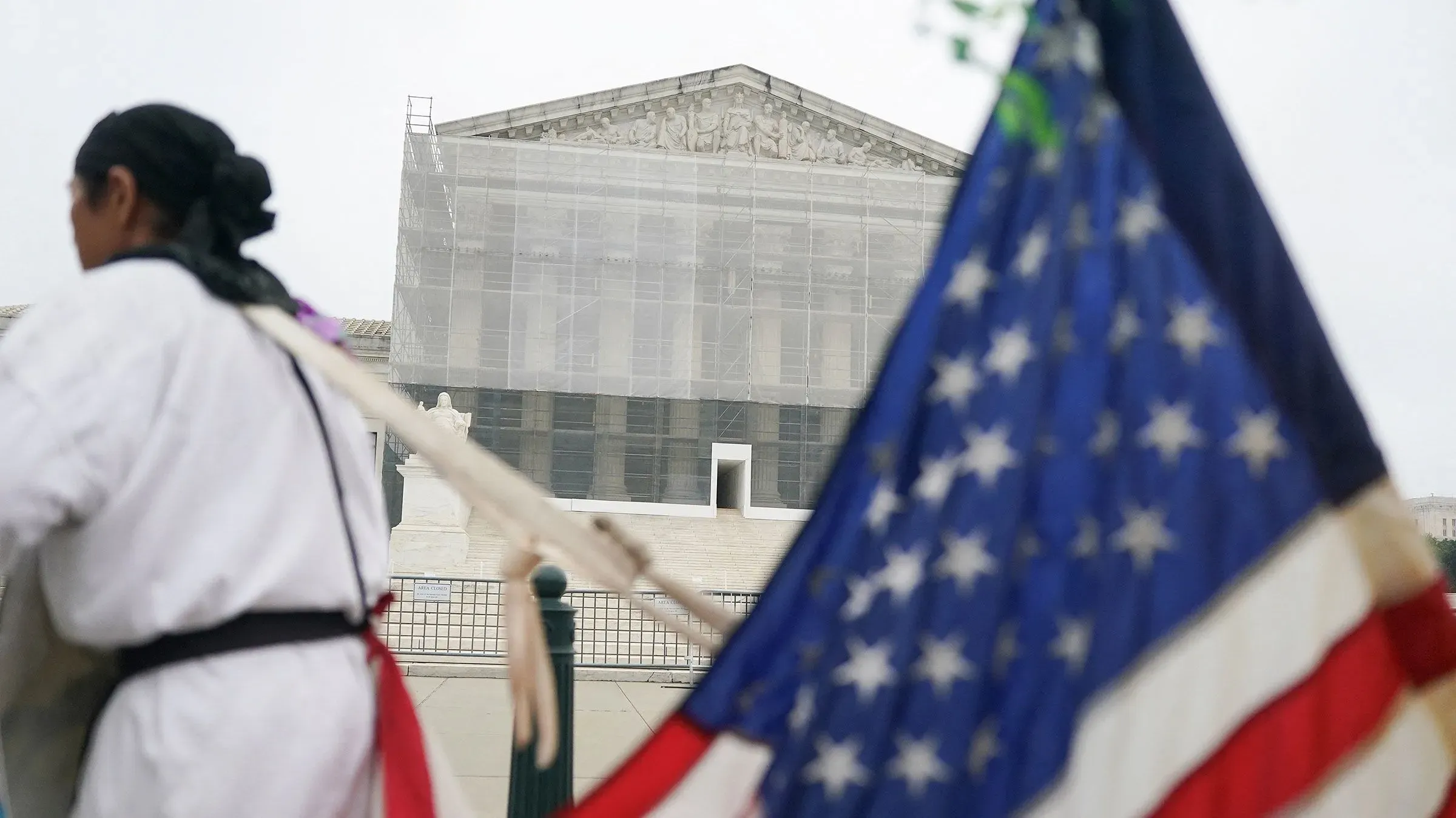 محتجون يقفون أمام المحكمة العليا الأمريكية، مع وجود علم أمريكي في المقدمة، بينما تُغطى واجهة المبنى بشبكة.