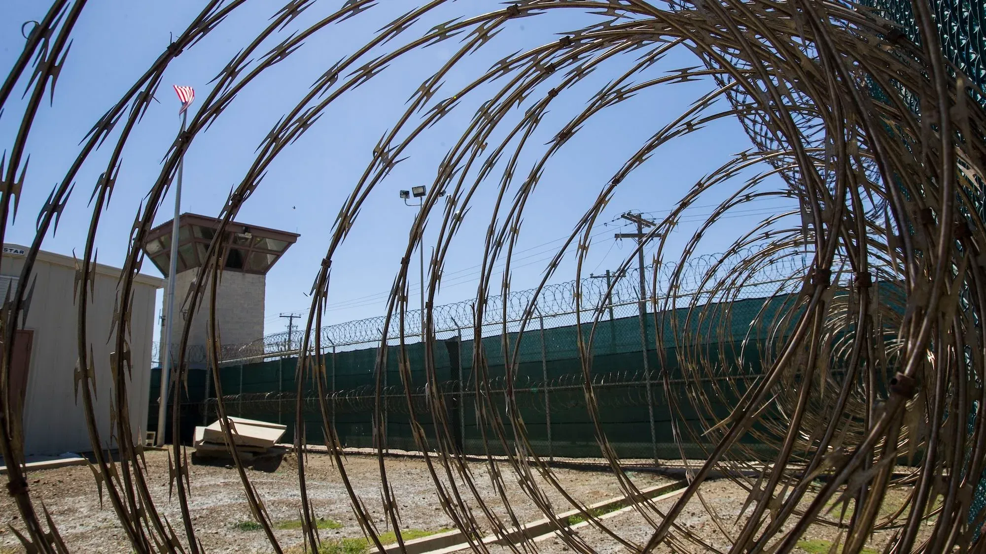 سياج شائك يحيط بقاعدة غوانتانامو البحرية، مع العلم الأمريكي يرفرف في الخلفية، يعكس التوترات السياسية حول قضايا الهجرة.