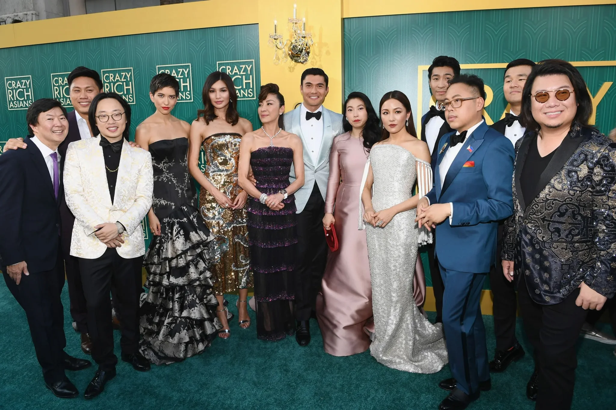 طاقم تمثيل فيلم "Crazy Rich Asians" يرتدون أزياء أنيقة في حفل عرض الفيلم، مع خلفية شعار الفيلم باللون الأخضر.
