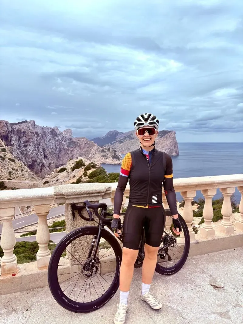امرأة مبتسمة ترتدي ملابس دراجات رياضية وتقف بجانب دراجتها الهوائية، مع منظر جبلي خلاب في الخلفية، تعكس روح التحدي في سباق فرنسا.