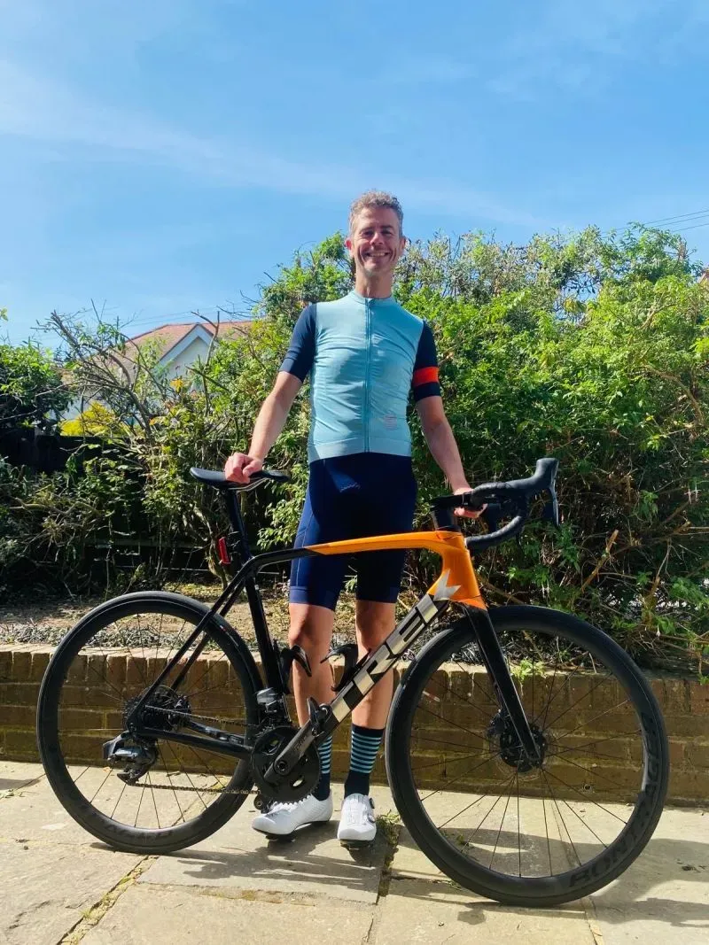 راكب دراجة مبتسم يرتدي ملابس رياضية، يقف بجانب دراجته في حديقة، مستعد للمشاركة في تحدي سباق طواف فرنسا للدراجات الهوائية.