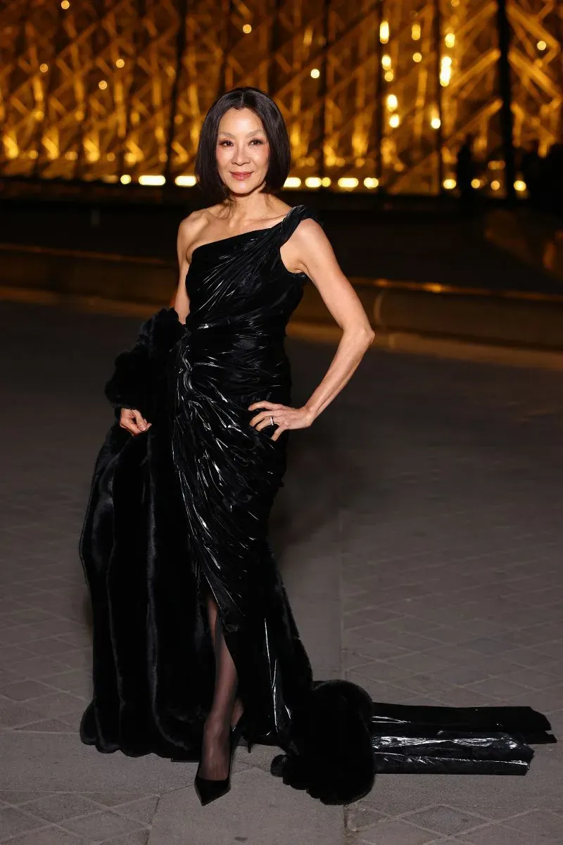 ممثلة ترتدي فستاناً أسود أنيقاً وتبتسم أمام متحف اللوفر، مع إضاءة خلفية تبرز الأجواء الفاخرة لحدث الموضة.