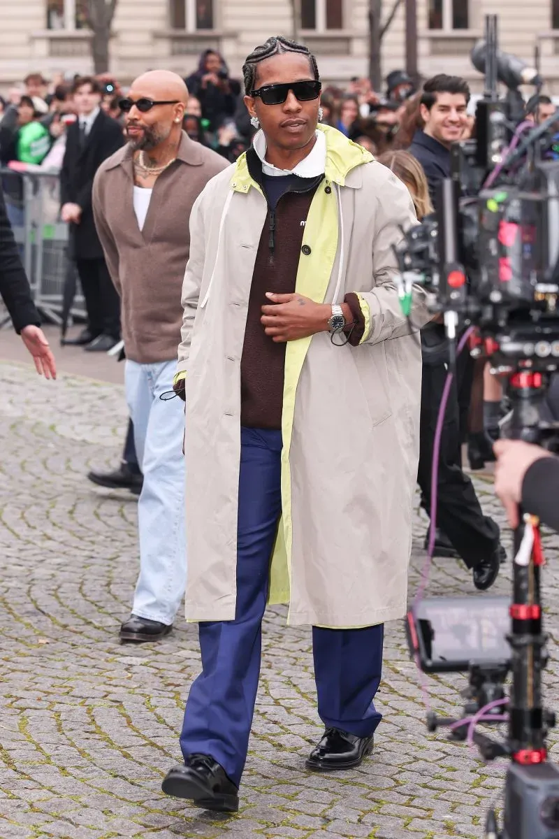 شخص يرتدي معطفًا خفيفًا بلونين مع بنطال أزرق، يتجول في الشارع أثناء أسبوع الموضة في باريس، محاطًا بجمهور ومصورين.