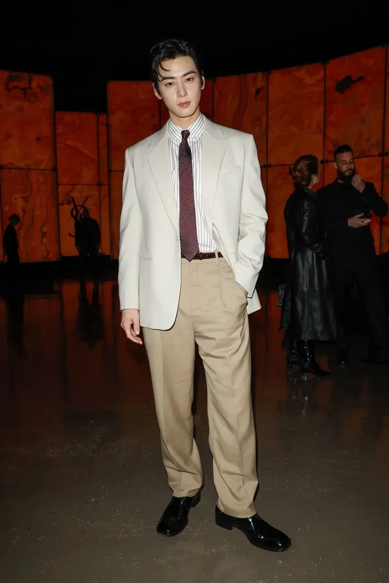 شاب يرتدي بدلة أنيقة باللونين الأبيض والبيج مع ربطة عنق بنية، يقف في عرض أزياء في باريس مع خلفية فنية.