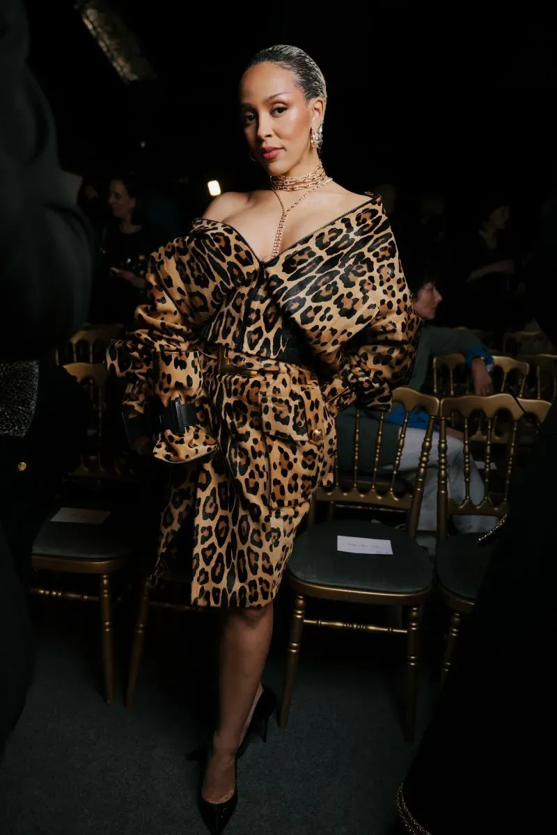 عارضة أزياء ترتدي فستاناً بنقشة النمر، تتوسط مجموعة من الكراسي في عرض أزياء، مع إضاءة خافتة تعكس أجواء الحدث.