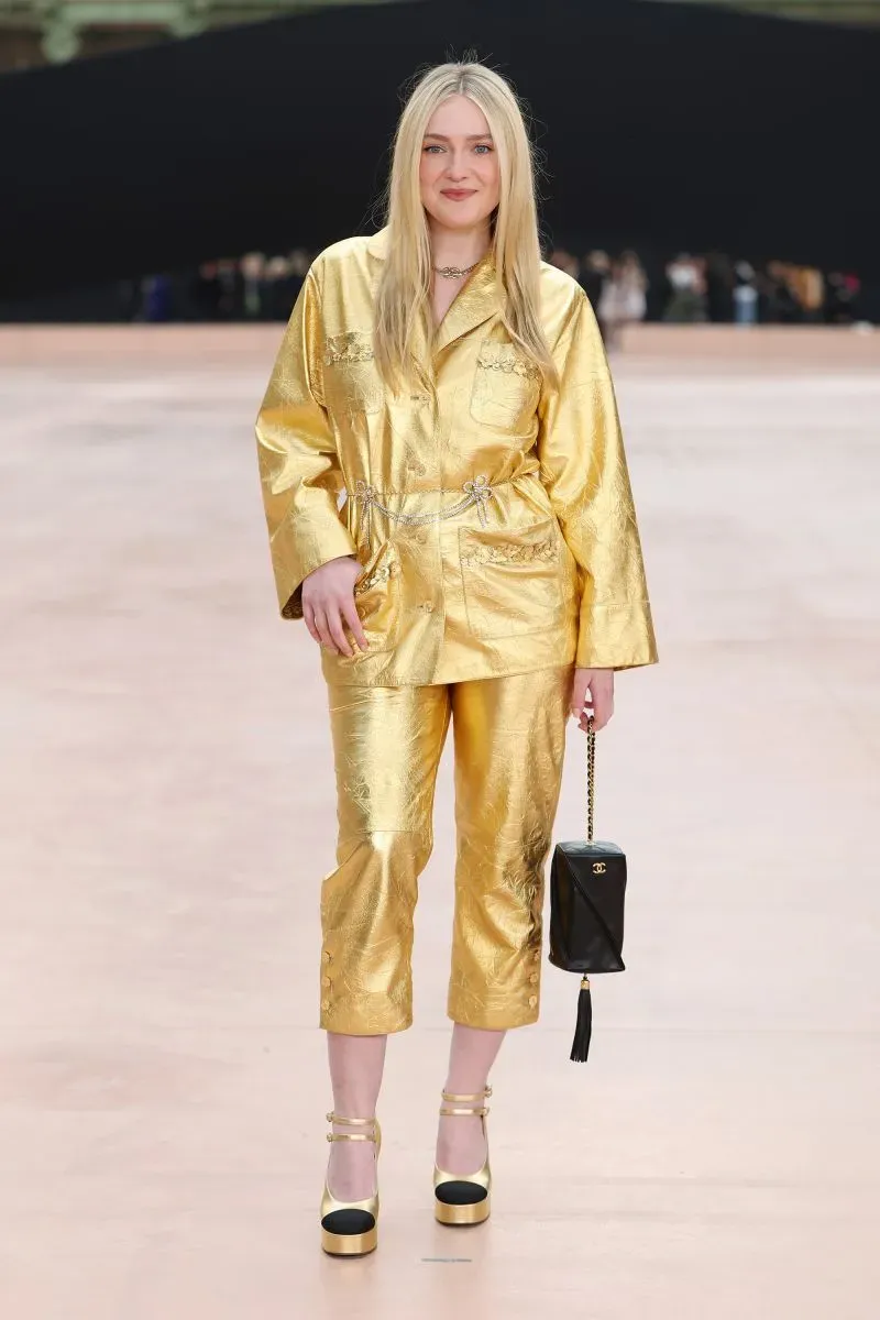 شابة ترتدي بدلة ذهبية لامعة مع حذاء بكعب عال، تحمل حقيبة صغيرة، وتبتسم في عرض أزياء باريس.