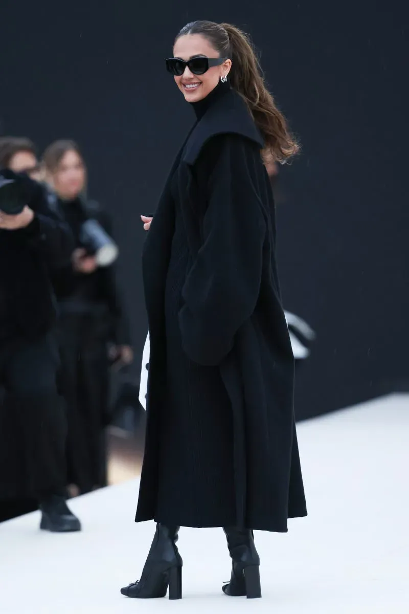 عارضة أزياء ترتدي معطفاً أسود طويلًا ونظارات شمسية، مبتسمة أثناء حضورها عرض أزياء في باريس.