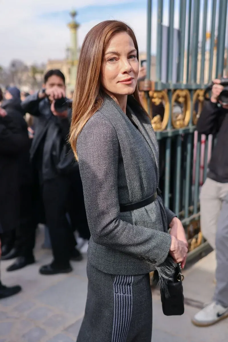 امرأة ترتدي بدلة أنيقة باللون الرمادي، تقف أمام بوابة متحف اللوفر مع مجموعة من المصورين في الخلفية، خلال أسبوع الموضة في باريس.