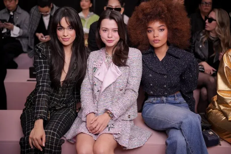 ثلاث نساء يجلسن في الصف الأمامي خلال أسبوع الموضة في باريس، يرتدين أزياء أنيقة مع تسريحات شعر مميزة، محاطات بالحضور.