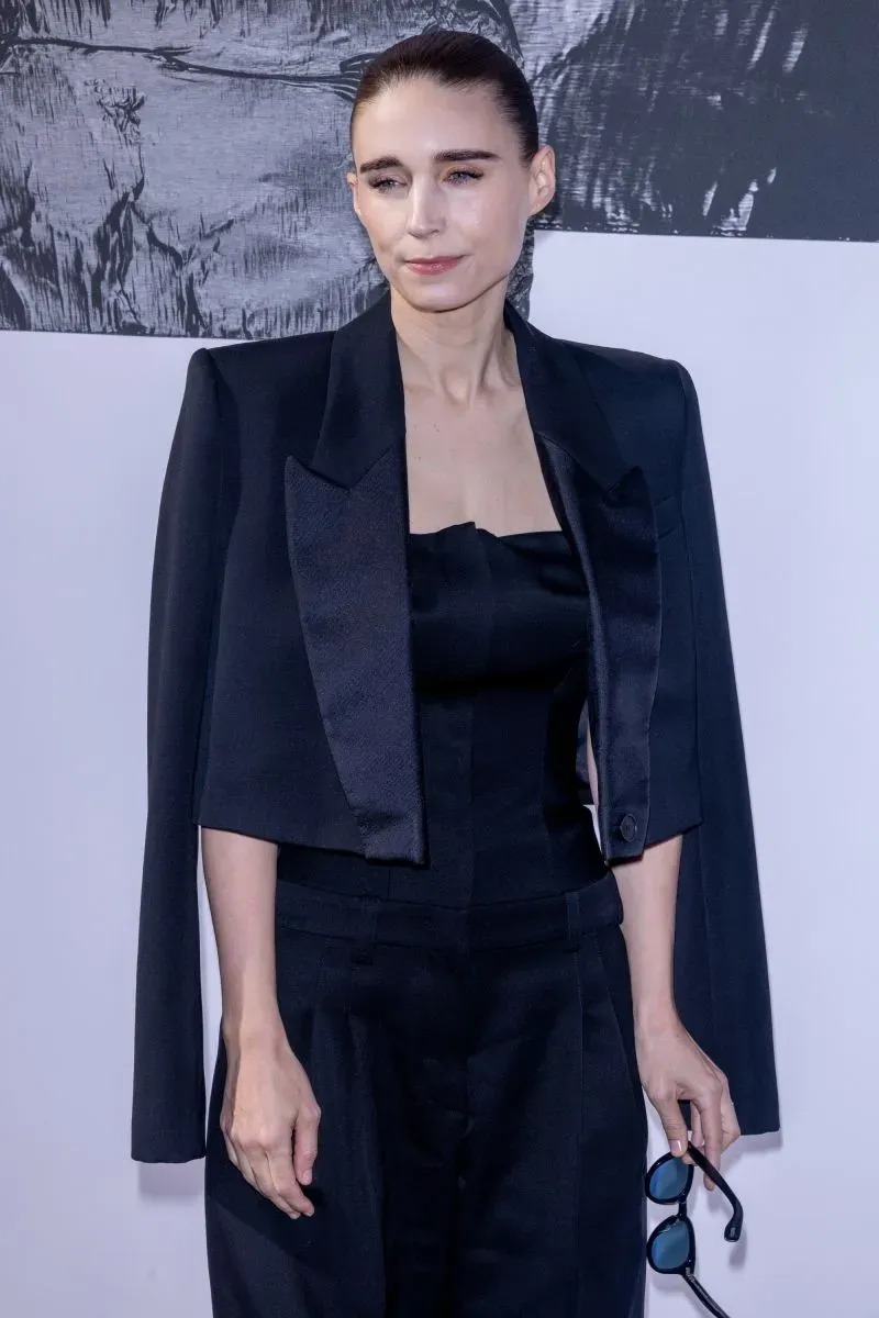 رووني مارا ترتدي بدلة سوداء أنيقة، تحمل نظارات شمسية، في عرض أزياء ضمن أسبوع الموضة في باريس لخريف وشتاء 2025.