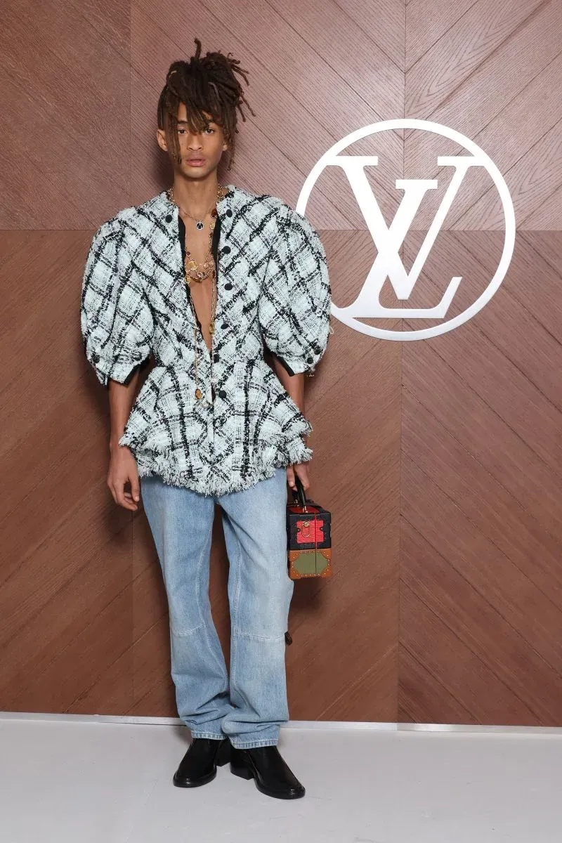 شاب يرتدي قميصًا مزخرفًا بأكمام منتفخة مع بنطال جينز، يحمل حقيبة صغيرة، يقف أمام شعار لويس فويتون في عرض أزياء باريس.