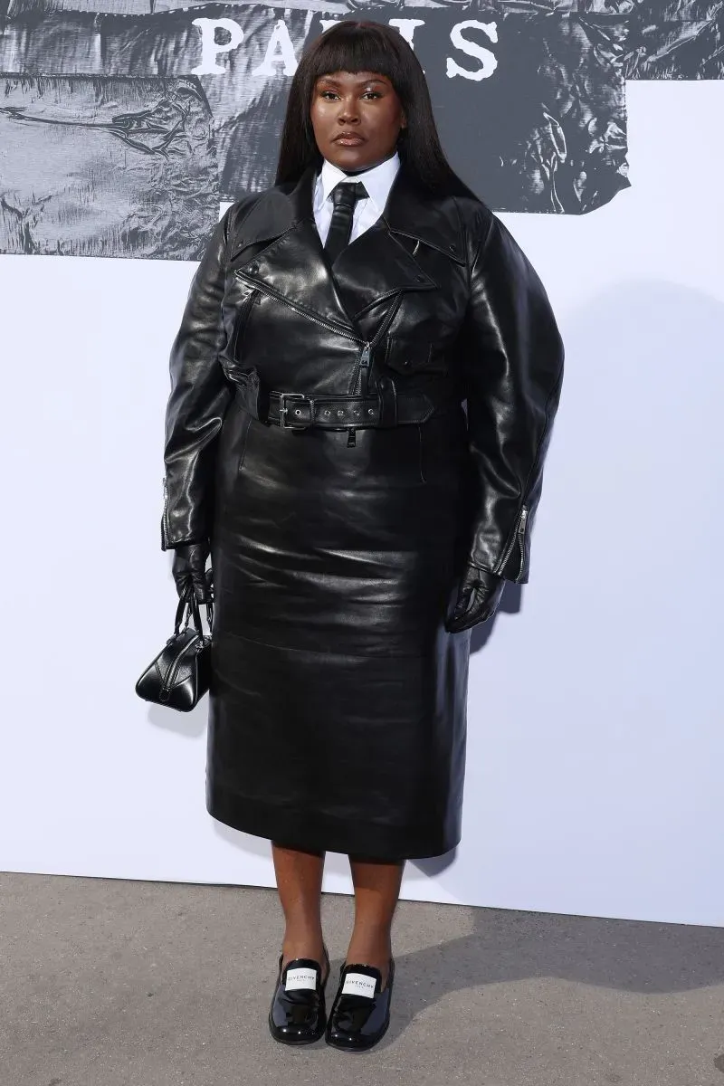 امرأة ترتدي فستاناً جلدياً أسوداً مع معطف طويل وقبعة، تحمل حقيبة صغيرة، في عرض أزياء باريس لخريف وشتاء 2025.