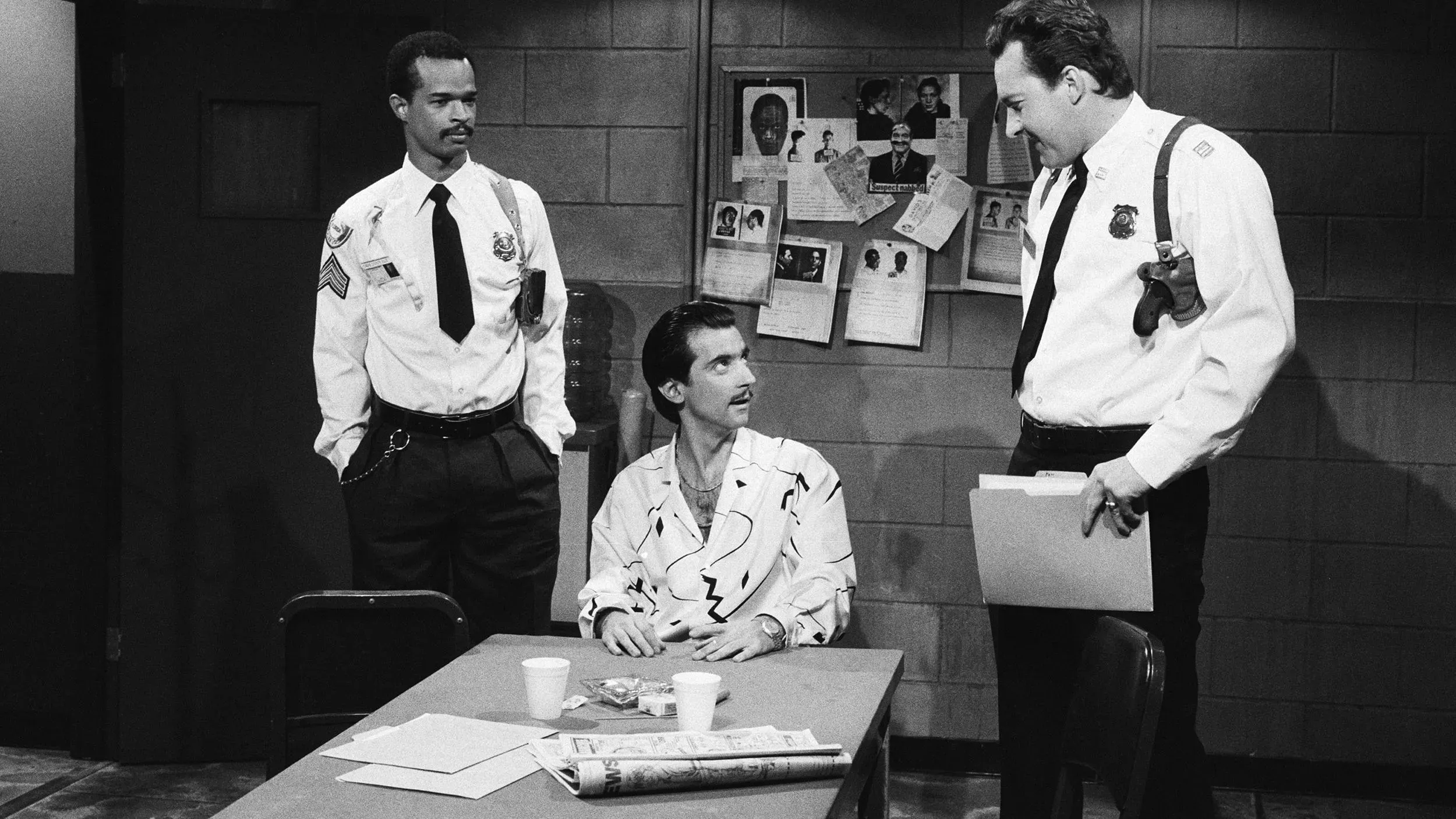 مشهد من برنامج "ساترداي نايت لايف" يظهر ثلاثة رجال في غرفة، مع رسومات وصور على الجدران، مما يبرز لحظة درامية من الكوميديا.