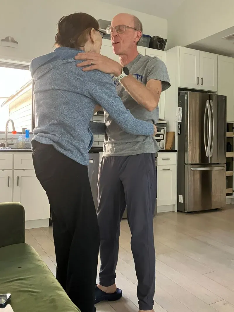 لحظة عاطفية بين مقدمي الرعاية في مطبخ منزلي، حيث يتبادلان الابتسامات والحنان، مع خلفية تعكس الحياة اليومية.