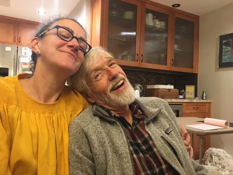 صورة تظهر امرأة مبتسمة تجلس بجانب رجل مسن ذو لحية بيضاء، في مطبخ دافئ. تعكس الصورة لحظات من السعادة والحميمية بينهما.