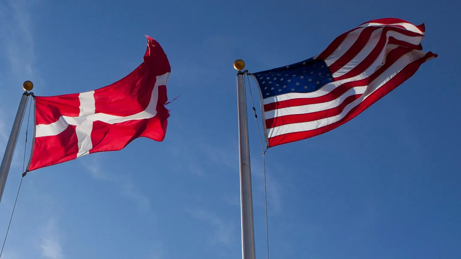 علم الدنمارك وعلم الولايات المتحدة يرفرفان معًا في السماء الزرقاء، مما يرمز إلى النقاش حول شراء كاليفورنيا.