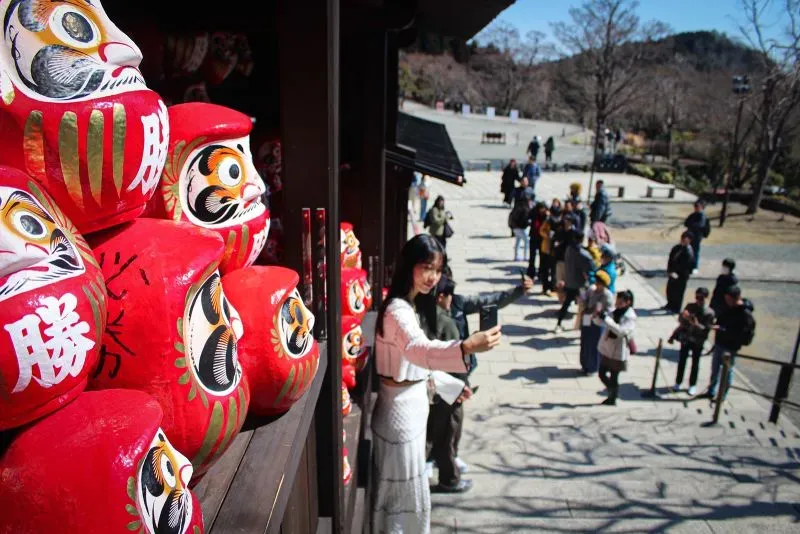 مجموعة من دمى داروما الحمراء في معبد كاتسو-جي، مع زائر يأخذ صورة سيلفي، تعكس رمزية الحظ والطموح في الثقافة اليابانية.