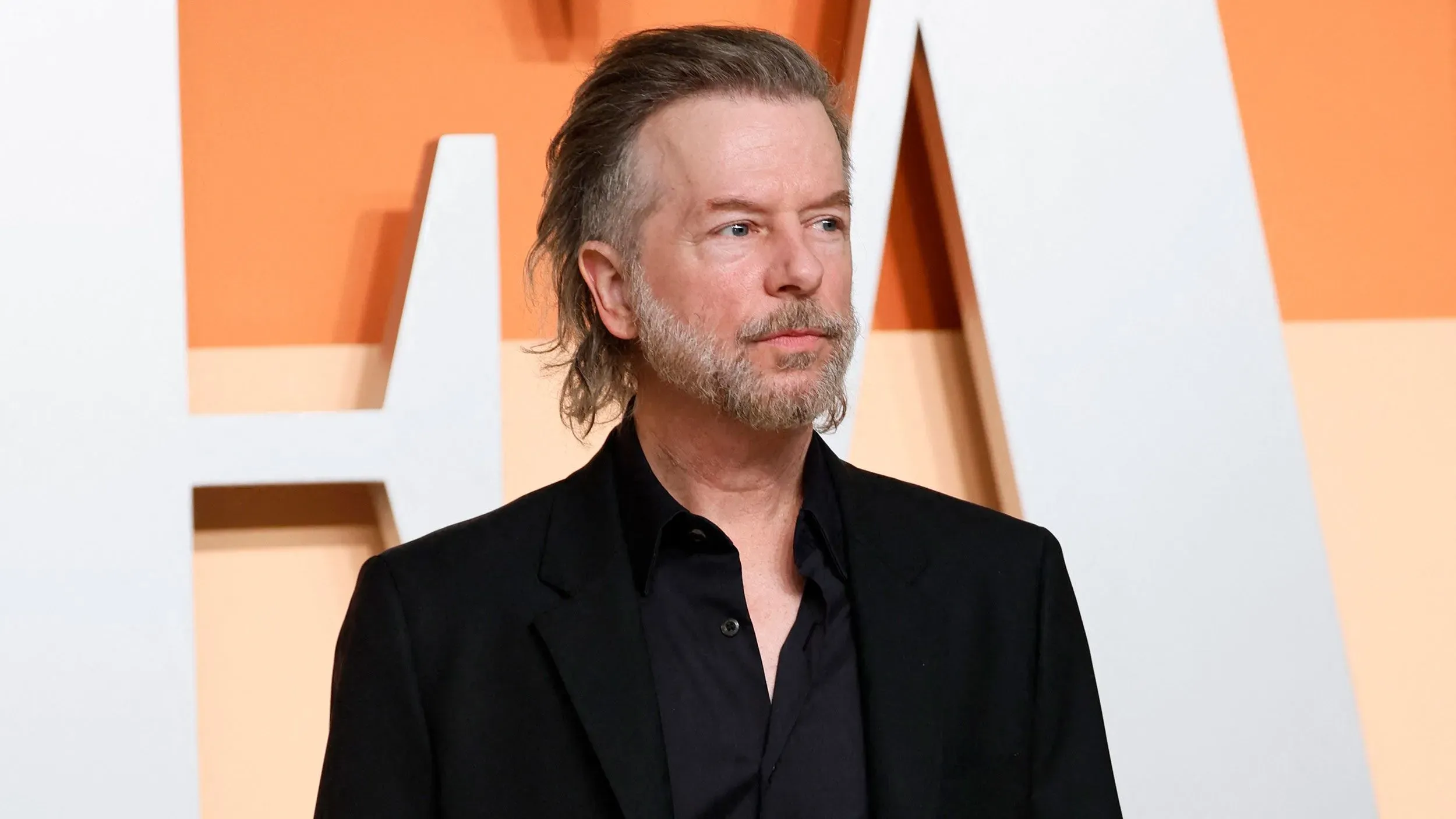 ديفيد سبيد يرتدي بدلة سوداء في حدث رسمي، مع خلفية برتقالية، يعبر عن قلقه بشأن موقف مخيف مع معجبيه.