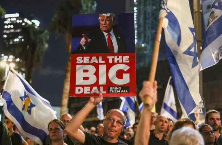 محتجون إسرائيليون يحملون لافتة تطالب بإبرام صفقة للإفراج عن الأسرى، مع أعلام إسرائيلية ترفرف في الخلفية خلال تظاهرة في تل أبيب.