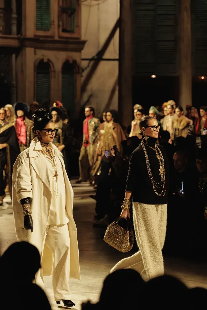 العرض يضم عارضتين تسيرتان على المنصة، تتزينان بأزياء فاخرة ومجوهرات متألقة، مع خلفية تعكس الطابع الثقافي لمومباي.