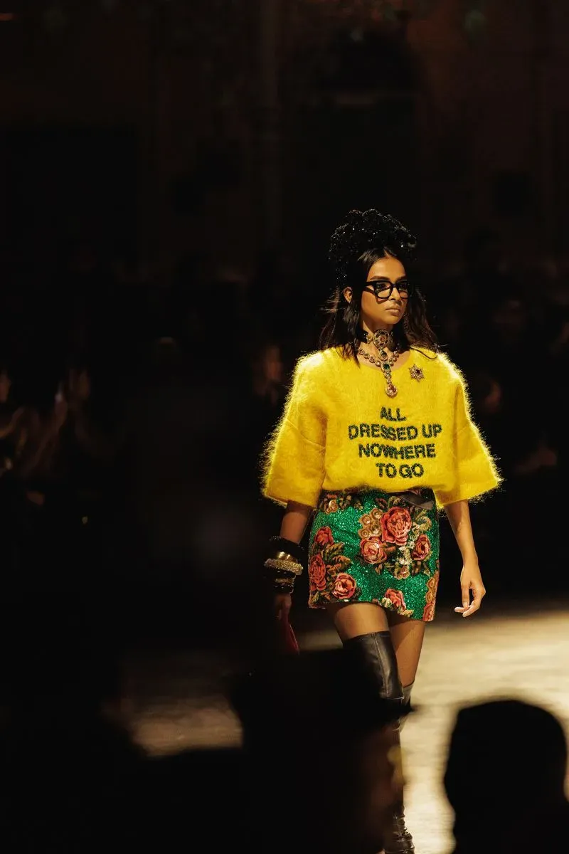 عارضة أزياء تسير على المنصة مرتدية سترة صفراء مع عبارة "ALL DRESSED UP, NOWHERE TO GO" وتنورة مزهرة، في عرض أزياء سابياساتشي موخرجي.