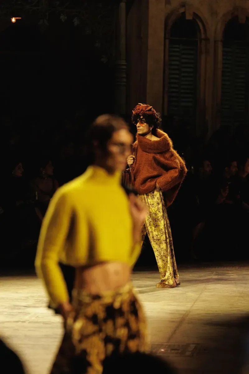 عارضة أزياء ترتدي كنزة بُنية وسروال مزخرف تسير على المنصة، بينما تظهر عارضة أخرى في الخلفية بلون أصفر، في عرض سابياساتشي موخرجي.