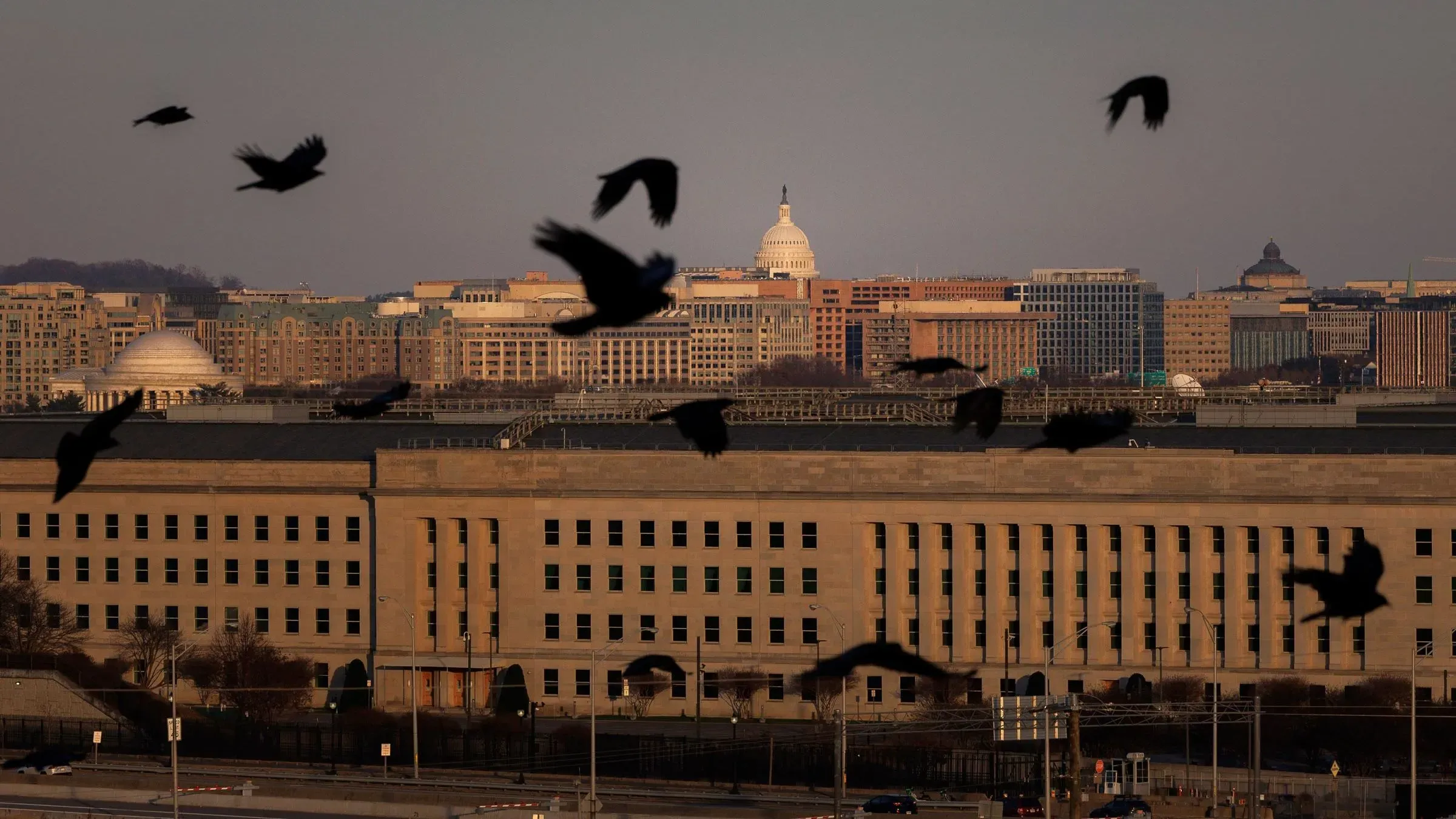 مبنى البنتاغون مع طيور تحلق في السماء، بينما تظهر معالم العاصمة واشنطن في الخلفية، مما يعكس أهمية القضايا العسكرية والأمنية.