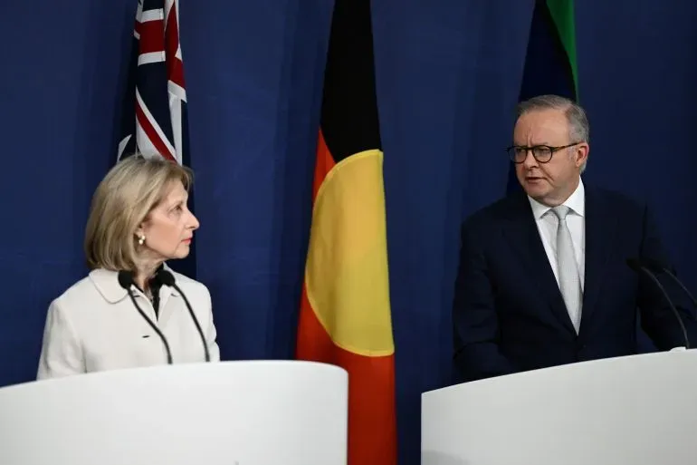 مبعوثة أسترالية لمكافحة معاداة السامية تتحدث مع رئيس الوزراء، بينما تلوح الأعلام الأسترالية والألمانية في الخلفية.