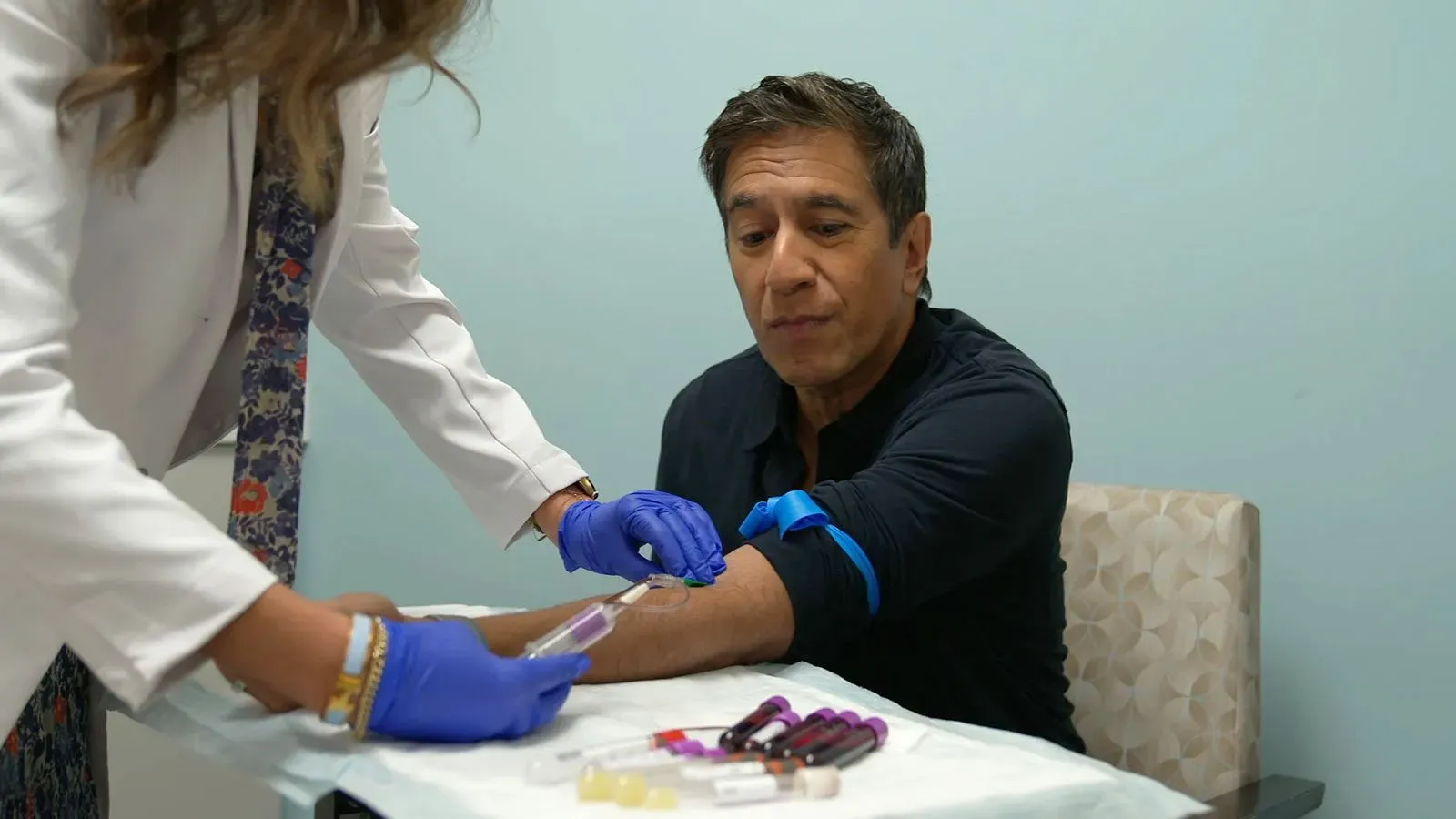 رجل يجلس في عيادة طبية أثناء سحب عينة دم، مع وجود عدة أنابيب اختبار ملونة على الطاولة، في سياق بحث حول الخرف.