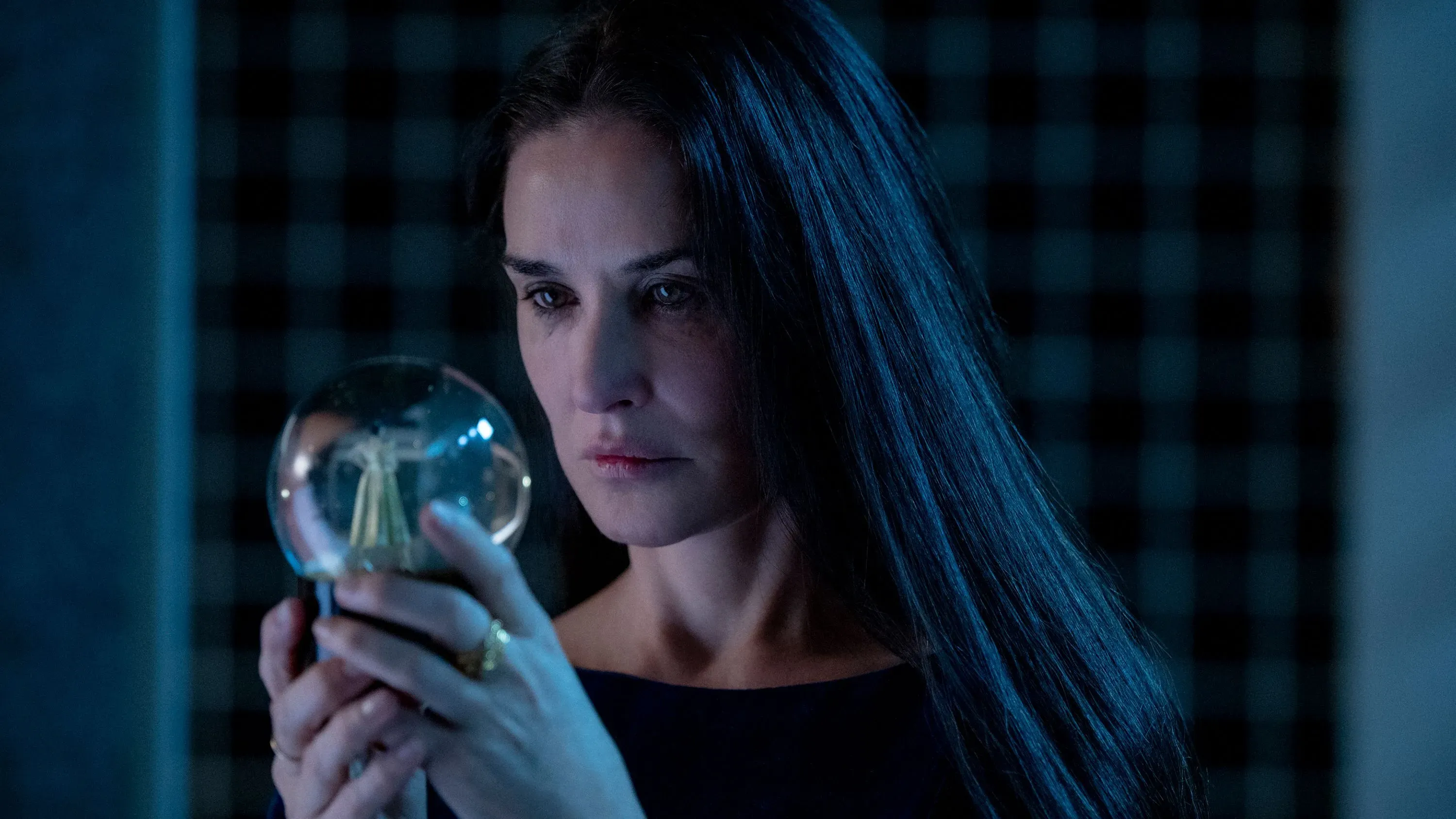 امرأة ذات شعر داكن طويل، تحمل كرة زجاجية في يدها، تعبر عن مشاعر عميقة في مشهد من فيلم "المادة" الذي يتناول قضايا كراهية النساء والتمييز.