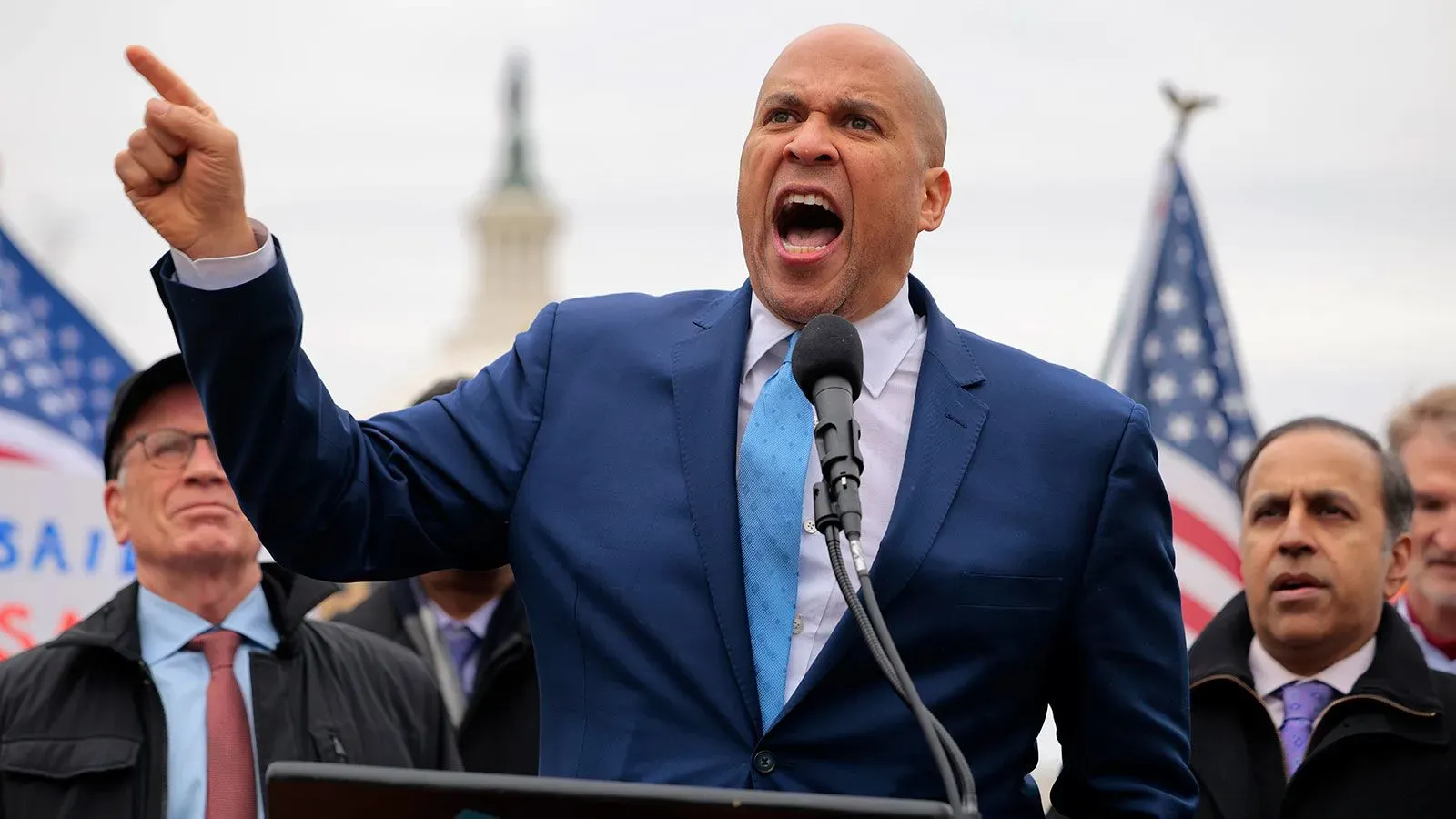 السناتور كوري بوكر يتحدث بحماس خلال خطاب احتجاجي في مجلس الشيوخ، مع خلفية من الأعلام الأمريكية، معبرًا عن قلقه بشأن أزمة البلاد.