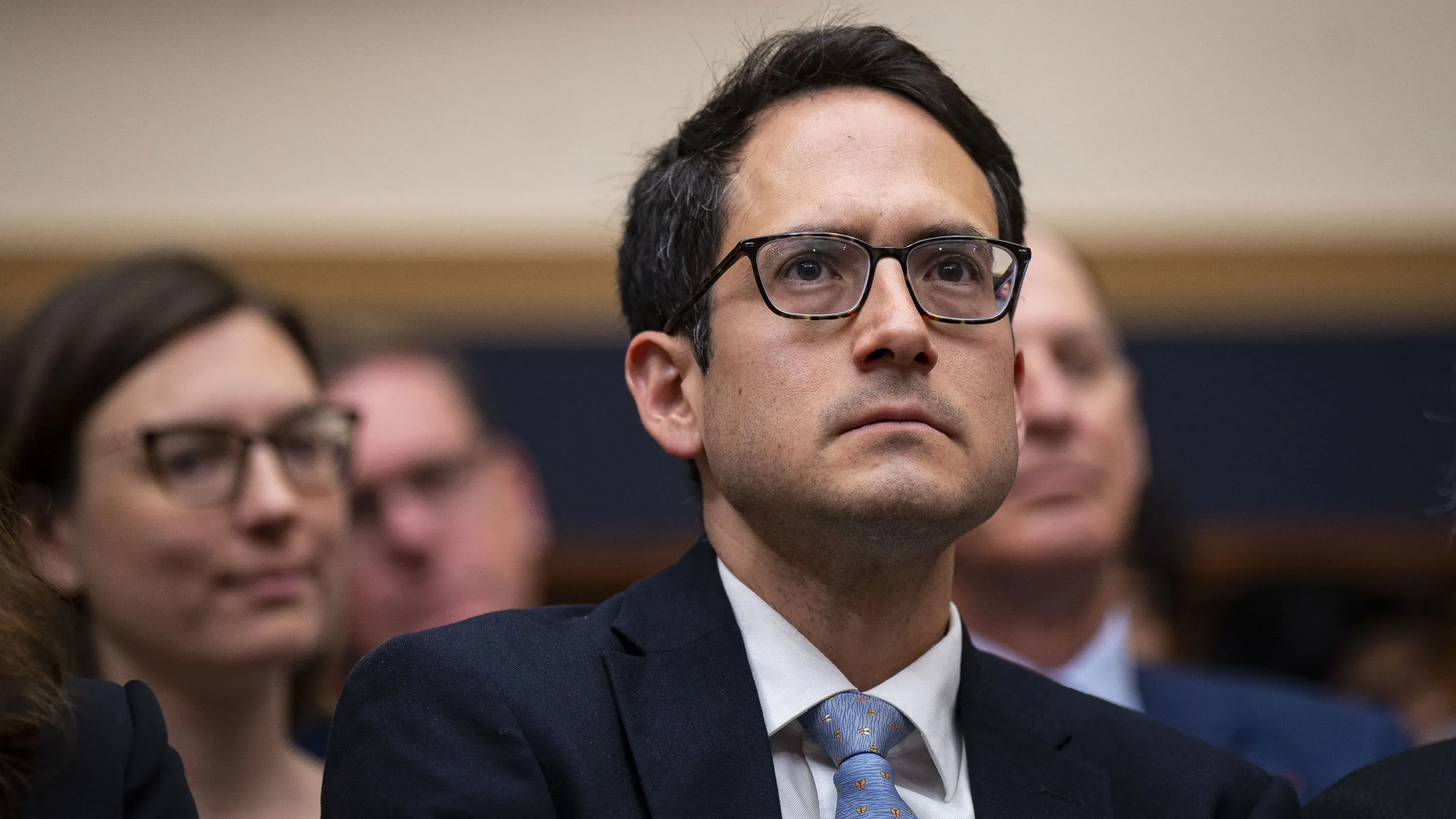 مجموعة من الأشخاص في جلسة استماع، مع تركيز على أحدهم الذي يبدو جادًا، مما يعكس الأجواء السياسية المتوترة حول إقالات ترامب للمفوضين.