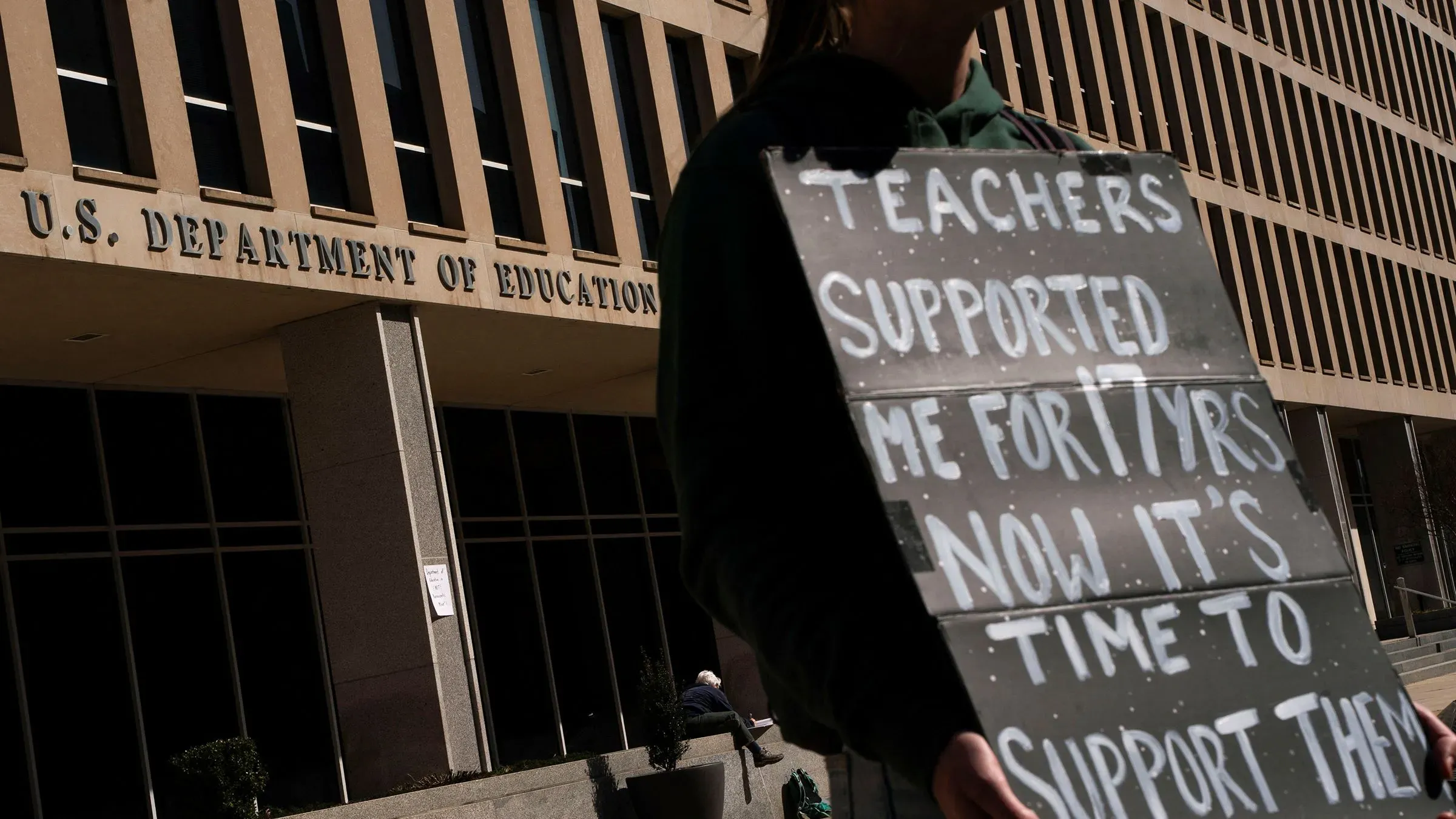 محتج يحمل لافتة أمام وزارة التعليم الأمريكية، يطالب بدعم المعلمين بعد تسريحات جماعية للموظفين، في سياق دعوى قضائية ضد الإدارة.