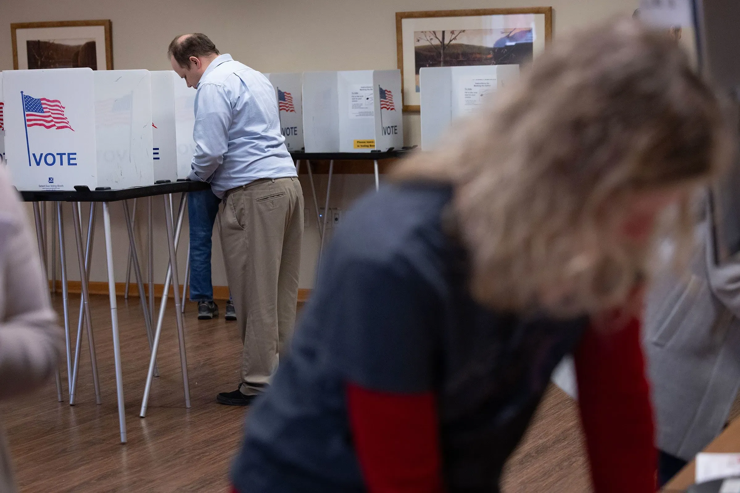 مواطنون يصوتون في مركز اقتراع، مع وجود صناديق اقتراع تحمل علم الولايات المتحدة، مما يعكس أهمية الانتخابات في البيئة السياسية الحالية.