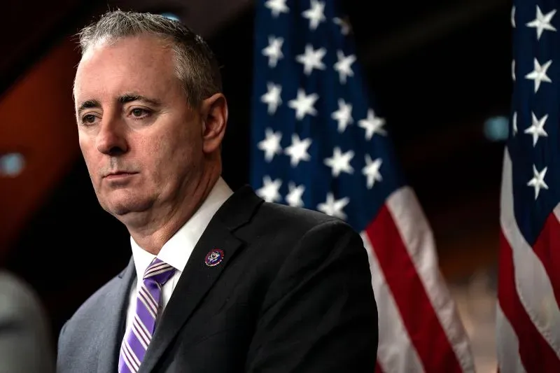 رجل يتحدث أمام علمين أمريكيين، معبرًا عن قضايا تتعلق بالسياسات الداخلية وقانون الهجرة وتأثيرها على المجتمع.